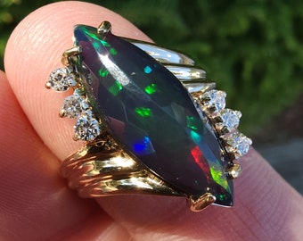 May include: A gold ring with a large, marquise-shaped black opal set in the center. The opal has a green, blue, and red fire. The ring is accented with smaller diamonds on either side of the opal.