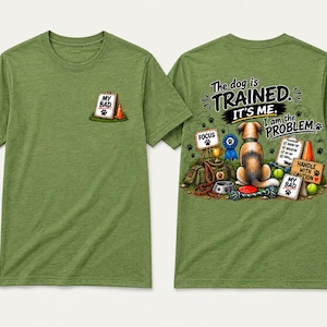 May include: An olive green t-shirt featuring a small graphic on the front left chest of a clipboard with the text MY BAD and an orange cone. The back displays a dog sitting with training gear, a blue ribbon, and text: The dog is TRAINED. IT'S ME. I am the PROBLEM.