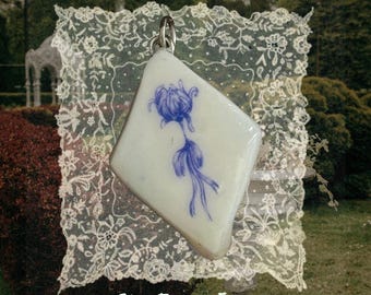 May include: A diamond-shaped white ceramic pendant featuring a hand-painted blue floral design. The pendant includes a silver-toned metal bail at the top for attachment. The text "She From Eden" is printed in white at the bottom of the image.