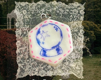 May include: A hexagonal white pendant featuring a blue ink illustration of a crystal ball with a star and sparkles. The border is decorated with small pink dashes. A silver-toned metal jump ring is attached to the top. The text She From Eden is visible.