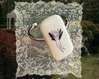 May include: A silver-toned metal cuff ring featuring a rectangular white ceramic centerpiece. The ceramic piece displays a small, dark, winged figure illustration. The text "She From Eden" is printed in white at the bottom of the image.