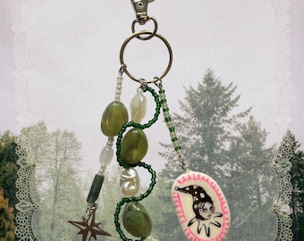 May include: A keychain featuring a silver-toned lobster clasp and ring, adorned with green and white beads, a dark star-shaped charm, and an oval pendant displaying a black and white illustration of a jester figure with pink painted edges.