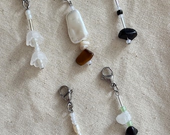 May include: Five handmade charm pendants with silver-tone clasps. The charms feature a variety of beads, including white flower-shaped beads, white and brown shell beads, and black and green stone beads. The pendants are displayed on a neutral fabric background.