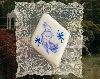 May include: A diamond-shaped white pendant featuring a blue ink illustration of a rabbit with a fish tail, flanked by two blue starburst symbols. The pendant includes a silver-toned metal jump ring at the top for attachment to a chain or cord.