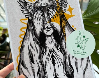 May include: A rectangular art print featuring a black and white ink illustration of a figure with wings on their head, covering their eyes with their hands, accented by yellow sunburst rays. A circular light green sticker shows two rabbits and the text She From Eden on etsy.com.