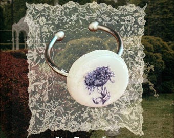 May include: A silver-toned metal cuff bracelet featuring a circular white ceramic centerpiece with a purple floral illustration. The text "She From Eden" is printed in white at the bottom of the image, which is layered over a square lace doily.