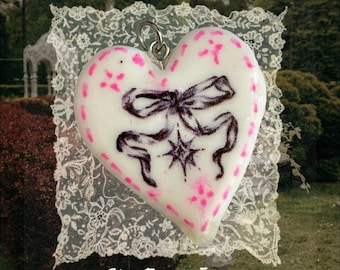 May include: A white heart-shaped pendant featuring a black ribbon bow illustration with a small star below it and pink dotted accents along the edges. The pendant includes a silver-toned metal bail at the top and is displayed over white lace.