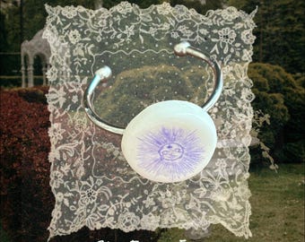 May include: A silver-toned metal cuff bracelet featuring a smooth, circular white stone centerpiece. The stone is decorated with a purple ink illustration of a sun with a face and radiating rays. The text She From Eden is printed below.
