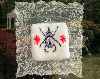 May include: A square, off-white pendant featuring a detailed black ink illustration of a beetle centered between two small, bright red star-like symbols. The pendant has a silver-toned metal jump ring at the top and is displayed against a white lace.