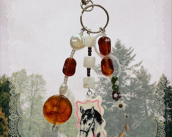 May include: A keychain featuring a silver-tone metal clasp and ring, adorned with three dangling strands of beads. The strands include amber-colored resin, white pearls, square white beads, and a central rectangular charm depicting a horse head.