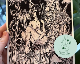 May include: Black and white art print featuring a seated figure with long hair, surrounded by flowers and mushrooms. The illustration has a detailed, hand-drawn style. A circular sticker with rabbit illustrations and text is visible.