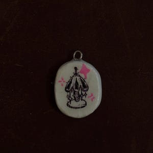 May include: A small, oval-shaped pendant with a silver-colored loop at the top. The pendant features a black illustration of a lamp with a decorative base and a pink star and starburst accents on a white background.