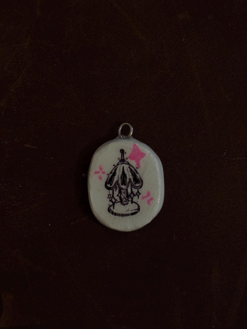 May include: A small, oval-shaped pendant with a silver-colored loop at the top. The pendant features a black illustration of a lamp with a decorative base and a pink star and starburst accents on a white background.