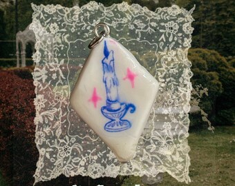 May include: A diamond-shaped white pendant featuring a blue illustration of a lit candle in a holder, accented by two small pink star shapes. The pendant includes a silver-toned metal jump ring at the top for attachment to a chain or cord.