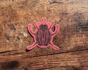 May include: A sticker featuring a stylized insect with a red body and dark brown, vertical striped markings on its back. The insect has six legs extending outward and a small head with two antennae, presented as a flat, die-cut graphic design.