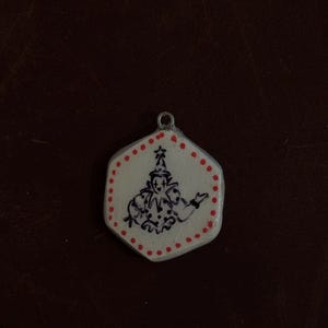 May include: A white, hexagonal pendant with a hand-drawn illustration of a jester in black ink. The pendant is outlined with red dots and a red star at the top. A small silver loop is at the top for hanging.