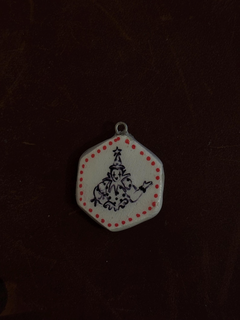 May include: A white, hexagonal pendant with a hand-drawn illustration of a jester in black ink. The pendant is outlined with red dots and a red star at the top. A small silver loop is at the top for hanging.