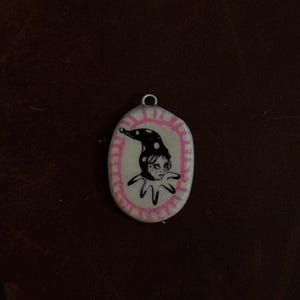 May include: Oval-shaped pendant with a pink border and a black and white jester illustration. The jester wears a polka-dot hat and a ruffled collar. The pendant has a silver loop at the top for hanging.