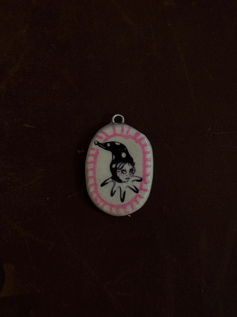 May include: Oval-shaped pendant with a pink border and a black and white jester illustration. The jester wears a polka-dot hat and a ruffled collar. The pendant has a silver loop at the top for hanging.