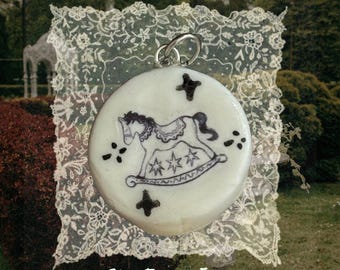 May include: A round, off-white ceramic pendant featuring a black line drawing of a rocking horse surrounded by small star and cross symbols. The pendant includes a silver-toned metal jump ring at the top and is displayed on a square lace doily.