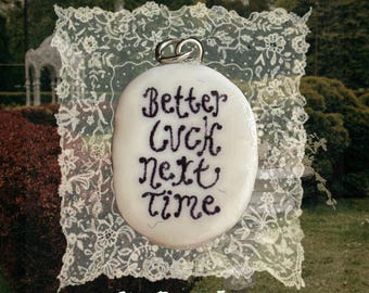 May include: An oval white pendant featuring the black embroidered text Better luck next time. The pendant is attached to a silver-toned metal jump ring and is displayed against a square piece of white lace fabric with a floral pattern.