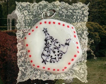 May include: A white, irregular hexagonal pendant featuring a black ink illustration of a clown wearing a pointed hat and ruffled collar. The edges are decorated with a series of small red dots. A silver-toned metal jump ring is attached at the top.
