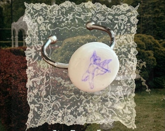 May include: A silver-toned metal cuff bracelet featuring a circular white ceramic centerpiece. The white surface displays a purple line drawing of a winged cherub holding a bow and arrow. The text "She From Eden" is printed in white at the bottom.