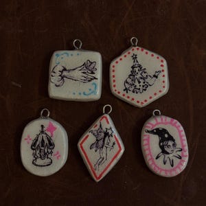 May include: A collection of six handmade pendants with unique illustrations. Each pendant features a different design, including a hand, a jester, and a fairy, all in a whimsical, vintage style. The pendants are white with black and pink accents, and have a metal loop at the top.