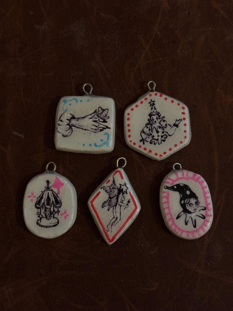 May include: A collection of six handmade pendants with unique illustrations. Each pendant features a different design, including a hand, a jester, and a fairy, all in a whimsical, vintage style. The pendants are white with black and pink accents, and have a metal loop at the top.