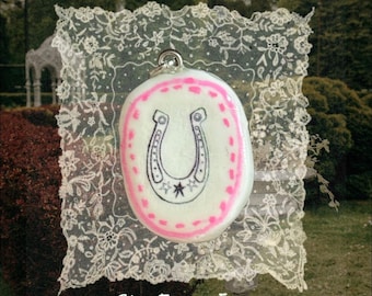 May include: A white oval pendant featuring a hand-drawn black horseshoe illustration with a small star at the base, surrounded by a pink painted border. The pendant includes a silver-toned metal bail at the top and is displayed on lace.