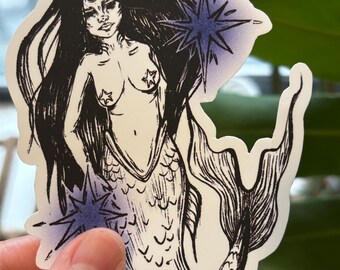 May include: A sticker featuring a black and white illustration of a mermaid with long flowing hair, a third eye, and star-shaped accents. The mermaid has a fish tail and is adorned with two star-shaped pasties. The sticker is held by a hand.