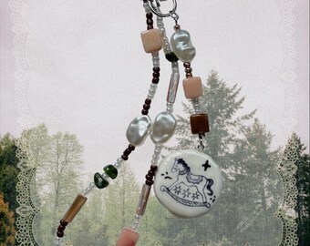 May include: A beaded keychain featuring a silver-toned lobster clasp, white irregular pearls, small dark brown beads, and square peach-colored beads. A round white ceramic charm displays a black line drawing of a rocking horse and a star.