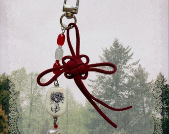 May include: A keychain featuring a silver lobster clasp, a red suede cord tied in a decorative knot, and a dangling charm strand. The strand includes red and white beads, a white oval pendant with a black floral illustration, and two white pearls.