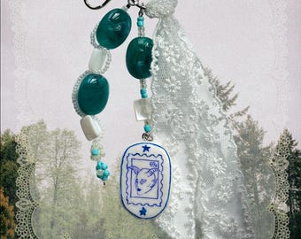 May include: A keychain featuring a silver metal ring and clasp, adorned with two strands of teal and white beads, a white lace ribbon, and a white oval charm with a blue stamp-style illustration of a horned figure and the text 69c.