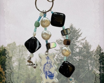 May include: A beaded keychain featuring a silver lobster clasp and ring, three strands of beads including black square beads, white pearls, and light blue accents, and a rectangular white charm with a blue illustration of a rabbit.