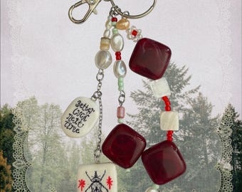 May include: A keychain featuring a silver metal ring and lobster clasp with three dangling strands. The strands include white and red beads, cream-colored pearls, two large dark red square beads, and two white tiles with black text and a beetle graphic.