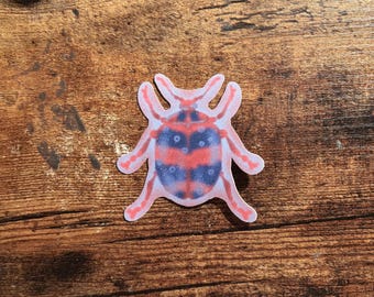May include: A sticker featuring a stylized beetle with a symmetrical pattern of alternating red and dark blue sections. The insect has six legs and two antennae, with a light-colored outline surrounding the entire shape of the beetle.