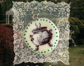 May include: A round, off-white pendant featuring a dark illustration of a three-legged cauldron with rising steam. The edge is decorated with a series of small green dots. A silver-toned metal jump ring is attached to the top of the pendant.