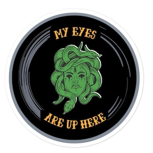 May include: A black and white sticker with a circular design. The center features a green illustration of a woman with snakes for hair. The text "MY EYES ARE UP HERE" is written in orange.