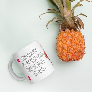 May include: White ceramic mug with the text "I CAN ALREADY TELL BY YOUR LAUGH THAT WE WON'T GET ALONG." The mug has red floral accents and is next to a ripe pineapple.