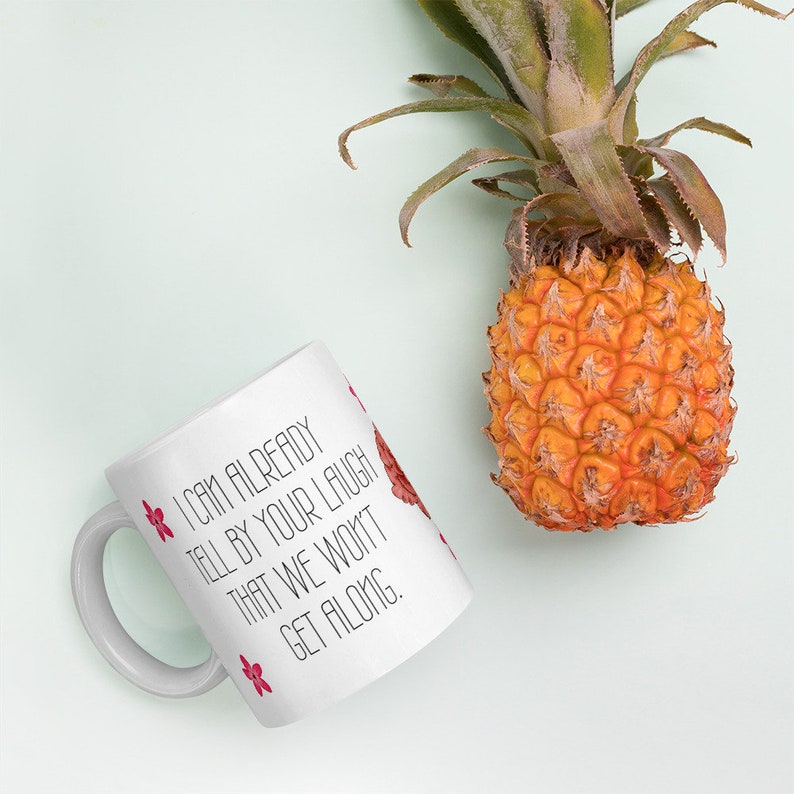 May include: White ceramic mug with the text "I CAN ALREADY TELL BY YOUR LAUGH THAT WE WON'T GET ALONG." The mug has red floral accents and is next to a ripe pineapple.