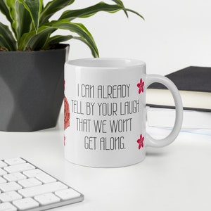 May include: White ceramic mug with the text "I can already tell by your laugh that we won't get along." The mug has a floral design and is next to a keyboard and a plant.
