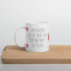 May include: White ceramic mug with a curved handle. The mug features the text "I CAN ALREADY TELL BY YOUR LAUGH THAT WE WON'T GET ALONG" in black, with red flower accents. The mug is on a wooden surface.