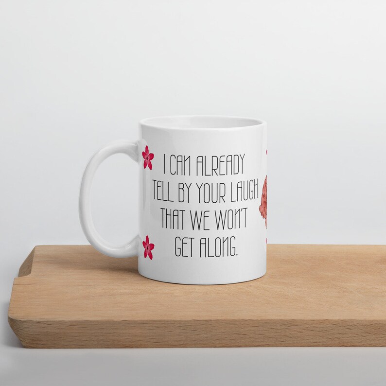 May include: White ceramic mug with a curved handle. The mug features the text "I CAN ALREADY TELL BY YOUR LAUGH THAT WE WON'T GET ALONG" in black, with red flower accents. The mug is on a wooden surface.