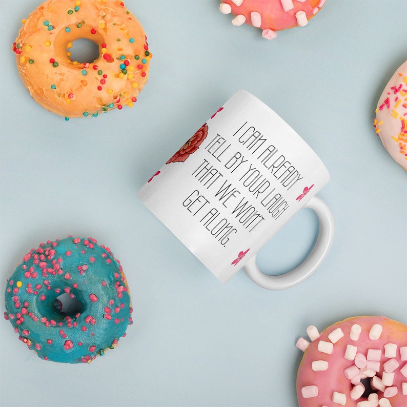 May include: White ceramic mug with the text "I CAN ALREADY TELL BY YOUR LAUGH THAT WE WON'T GET ALONG." The mug is surrounded by colorful donuts with sprinkles on a light blue surface.