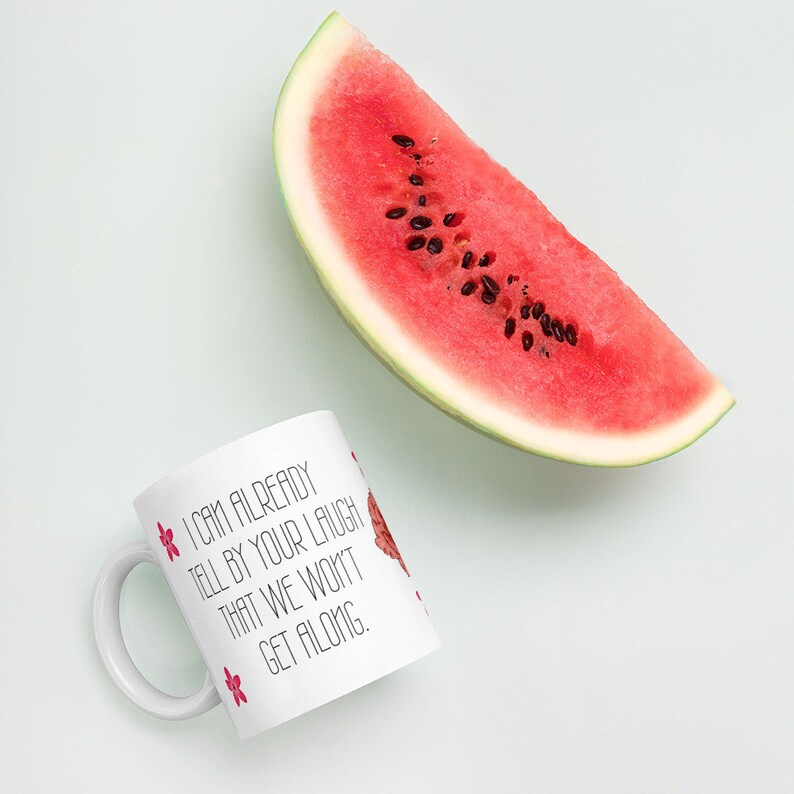 May include: A white ceramic mug with the text "I CAN ALREADY TELL BY YOUR LAUGH THAT WE WON'T GET ALONG." is decorated with pink flowers and a watermelon slice illustration. A fresh watermelon slice is also in the image.