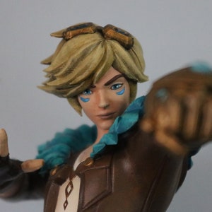 May include: A detailed figurine of a character with blonde hair, blue eye markings, and goggles. The figure wears a brown jacket with a teal collar and is posed with a clenched fist. The overall design suggests a fantasy or gaming theme.