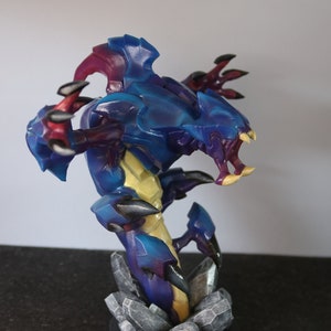 May include: A detailed figurine of a creature with a blue, purple, and yellow color scheme. The figure has sharp claws and teeth, and is mounted on a base of gray rocks. The figurine is approximately 8 inches tall.
