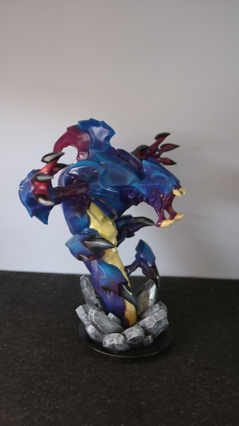 May include: A detailed figurine of a creature with a blue, purple, and yellow color scheme. The figure has sharp claws and teeth, and is mounted on a base of gray rocks. The figurine is approximately 8 inches tall.