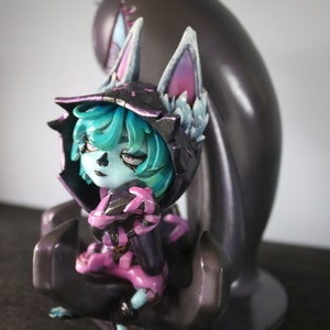 May include: A detailed figurine of a character with teal hair, a black hooded cloak, and pink accents. The figure is seated in a stylized pose, with a dark, amorphous shape behind it. The figurine is on a dark surface.